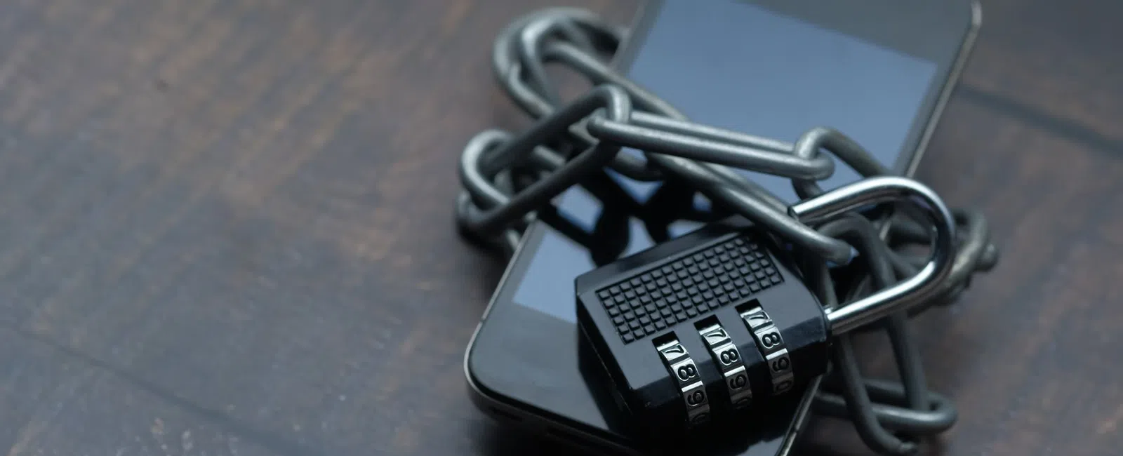 Smartphone wrapped in metal chain secured with combination padlock on wooden surface representing security.