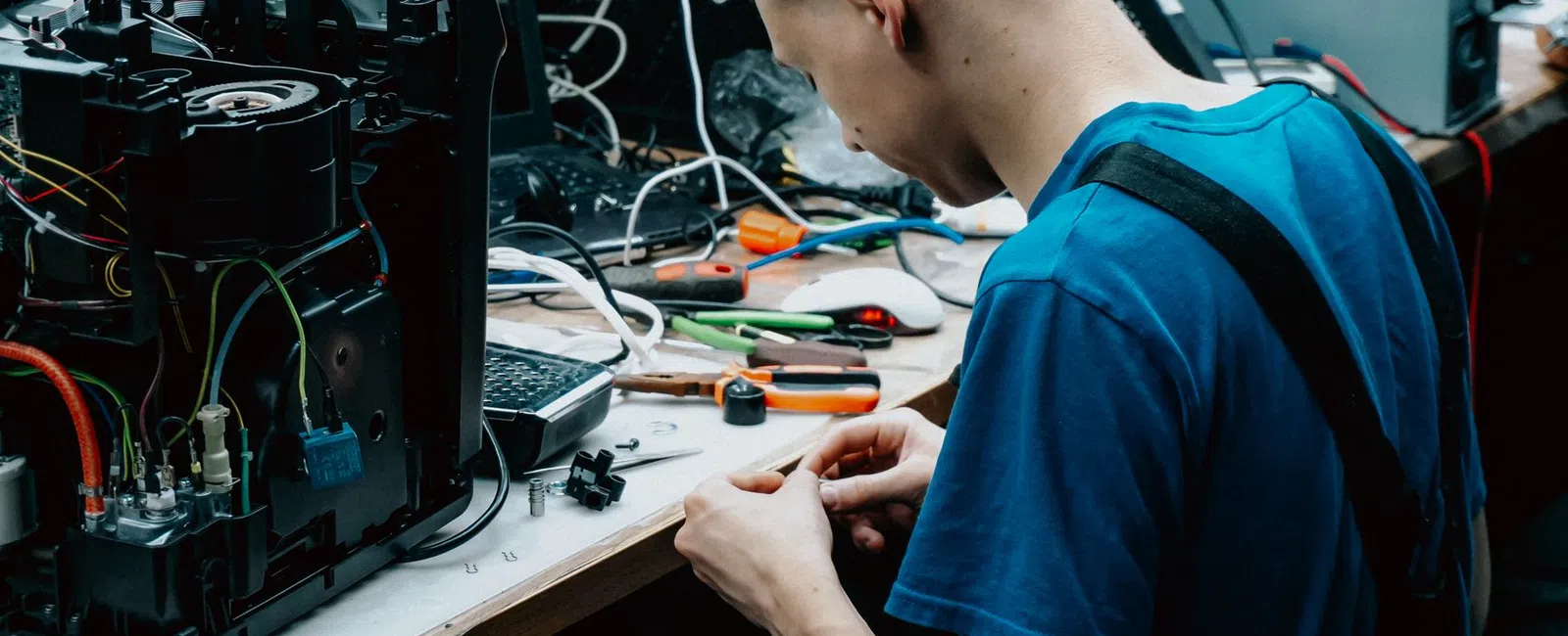 Technician repairing electronic device with wires and tools on a cluttered workbench in a workshop.