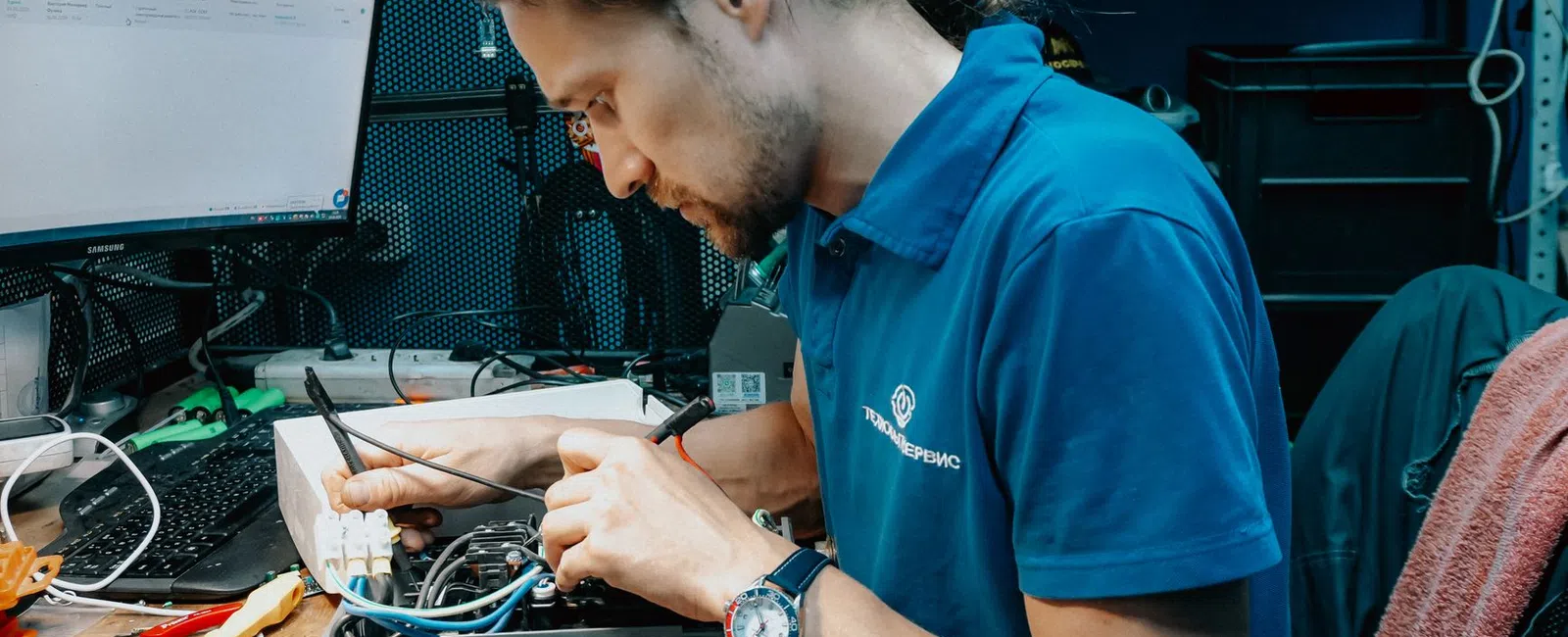 Technician repairing electronic device, testing circuit with multimeter at a cluttered workstation with computer screen.