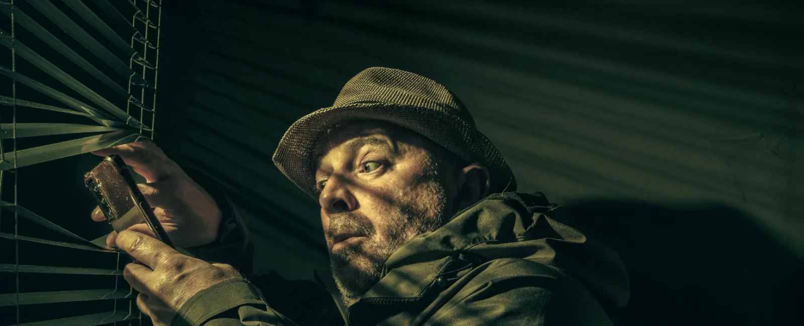Man in a hat and jacket peering through window blinds holding a phone in a dimly lit room at night.
