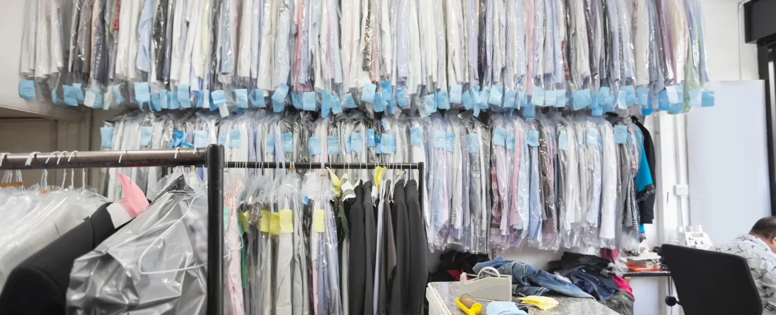 Interior of a busy dry cleaning shop with clothes hanging on racks covered in plastic and a cluttered counter area.