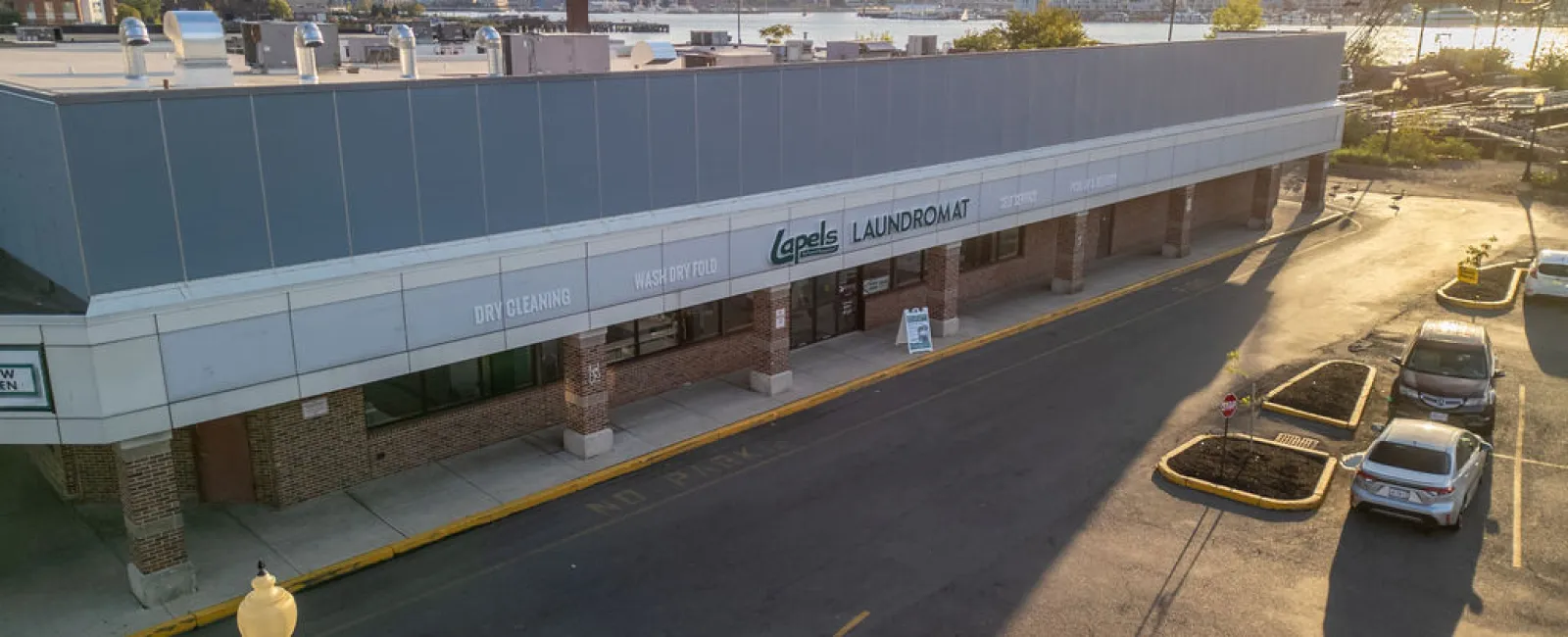 Laundromat storefront with dry cleaning and wash/dry/fold services, city skyline and river in background at sunset.