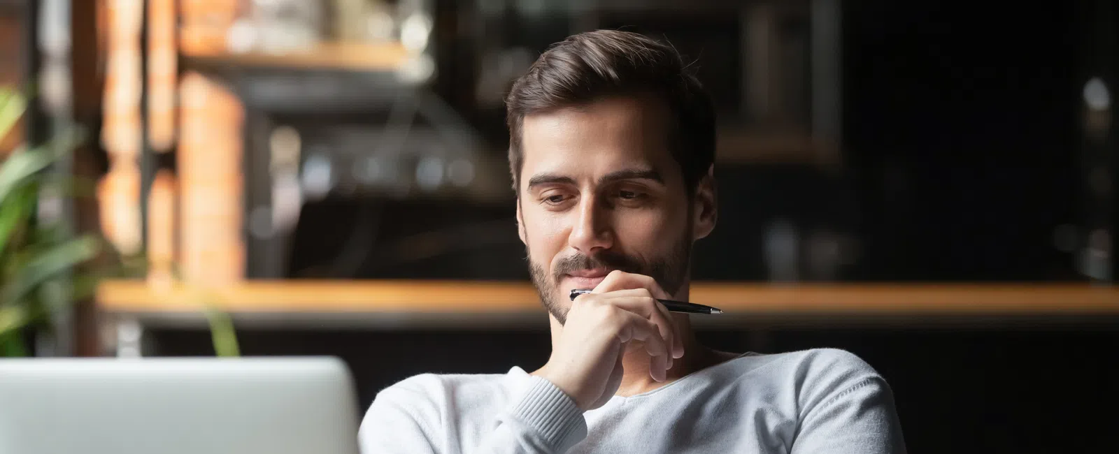 Man in white sweater thoughtfully working on laptop at cozy workspace with coffee mug and notes.