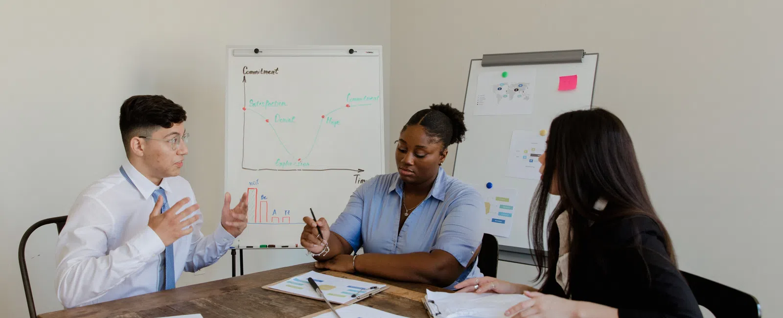 Three colleagues in a meeting discussing charts and graphs in a modern office setting with whiteboards.