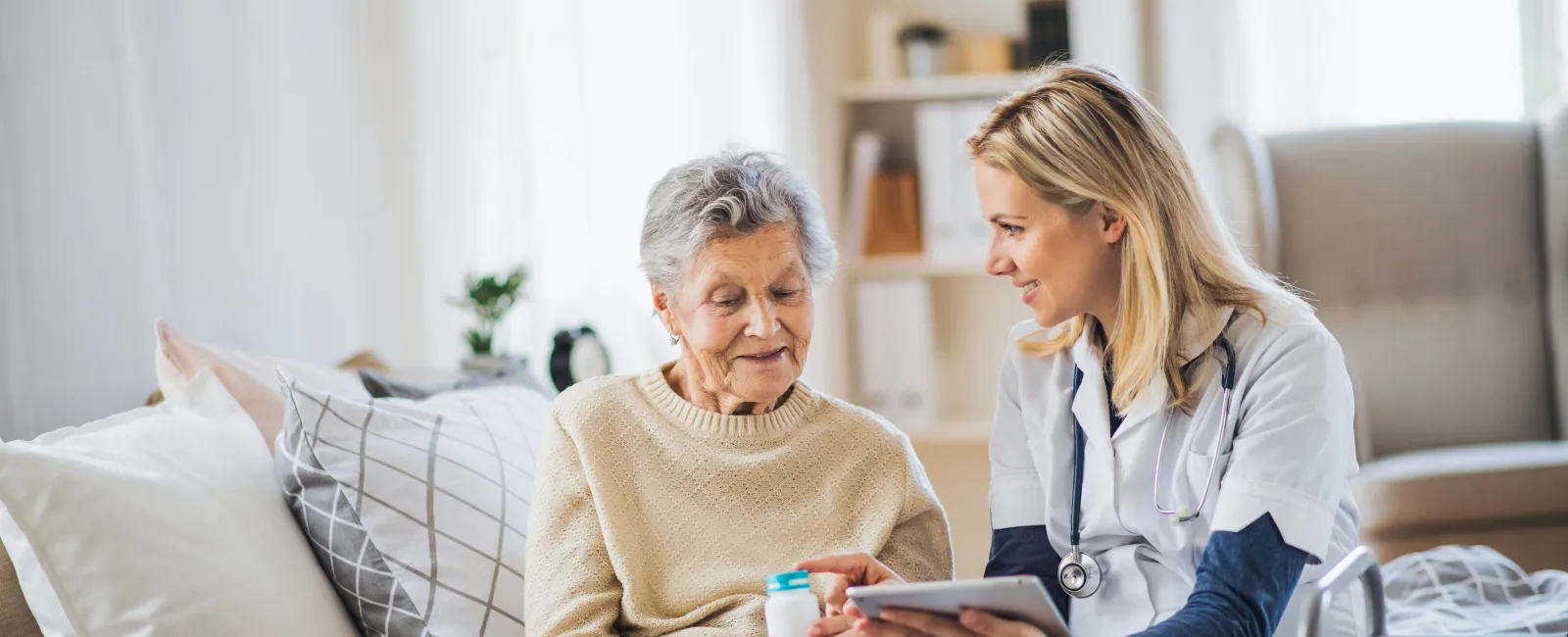 Senior woman receiving medical care from a female home healthcare provider holding a pill bottle and a tablet.