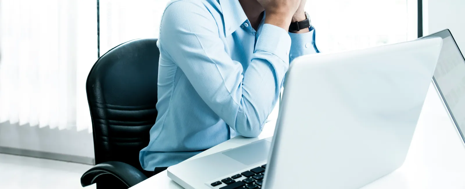 Stressed man in blue shirt with glasses sitting at desk covering face in front of laptop in real estate office in New Jersey.