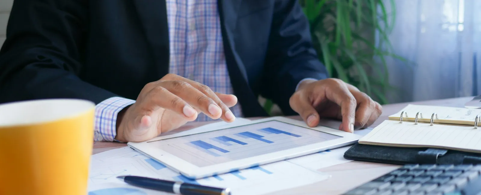 Business professional analyzing bar charts on a tablet at desk with calculator, pen, notebook, and coffee mug