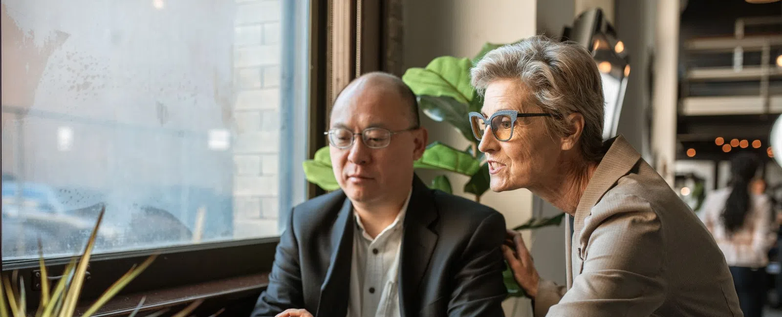 Two professionals collaborate on a laptop near a window in a modern office setting with plants and coffee.