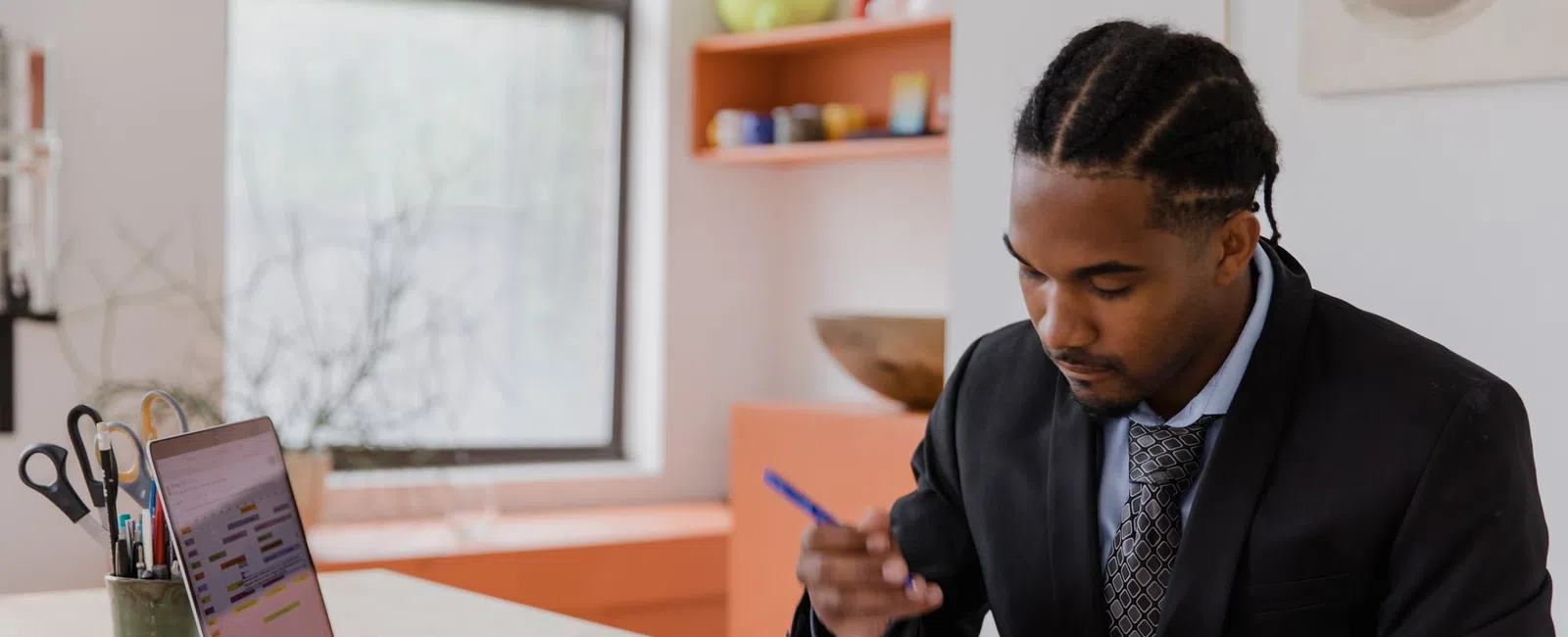 Professional man in suit reviewing documents at desk with laptop and office supplies near bright window.