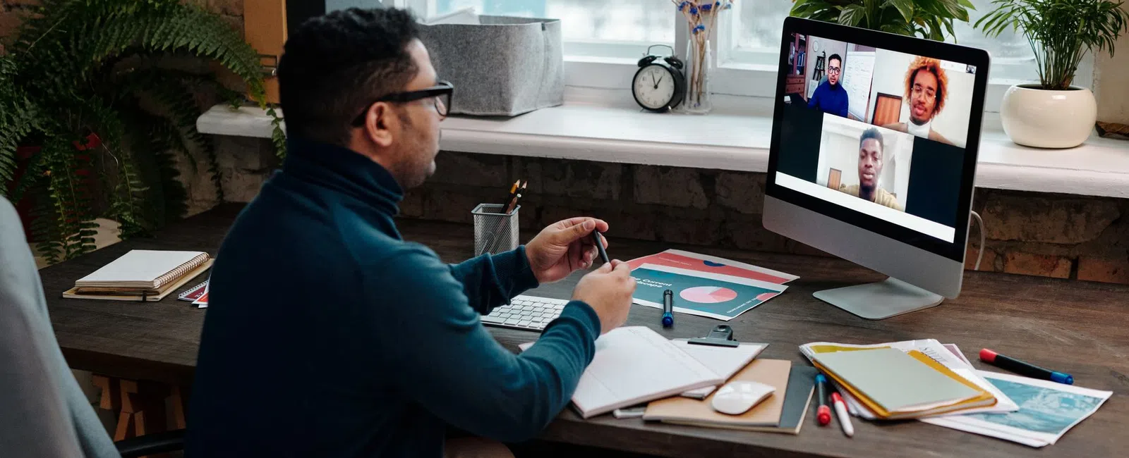 Man attending a virtual meeting on desktop computer in a home office with notebooks and plants around.
