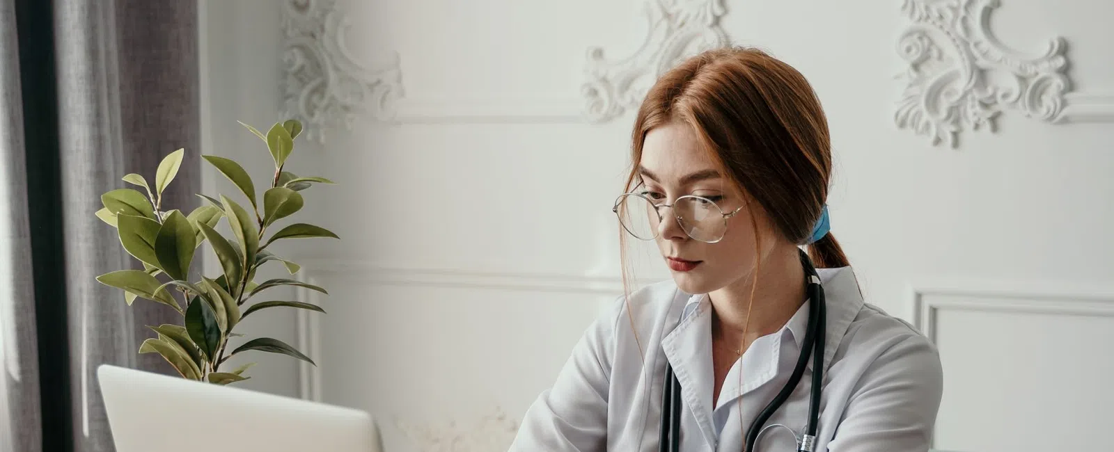 Female doctor with stethoscope working on laptop at desk with notebook and pills nearby