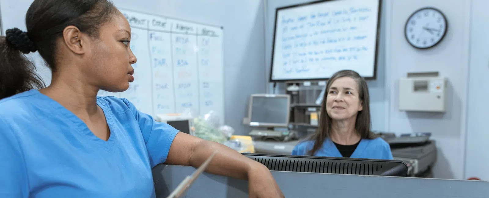 Two medical professionals in blue scrubs talking in a hospital nurse station with computer and whiteboard.
