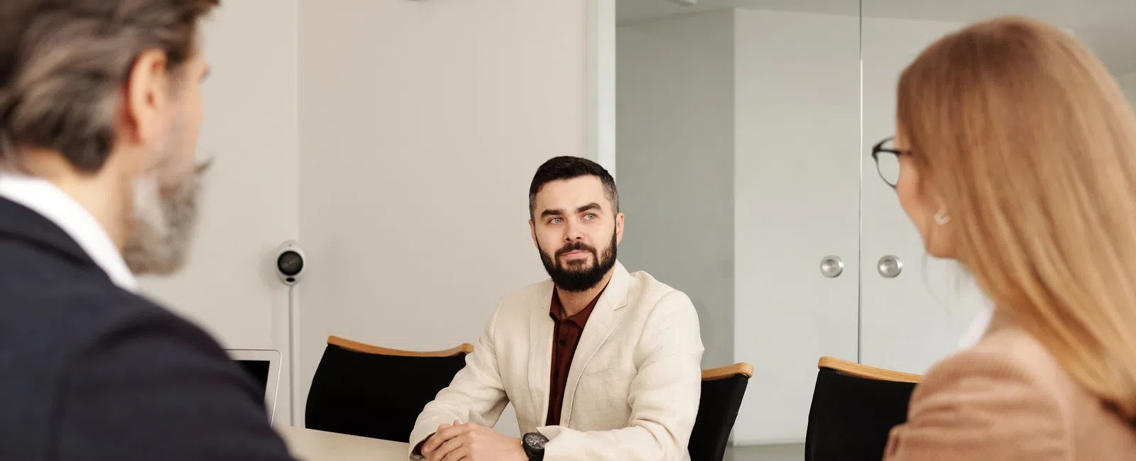 Man in beige blazer sitting at conference table engaged in discussion with two colleagues in office setting.