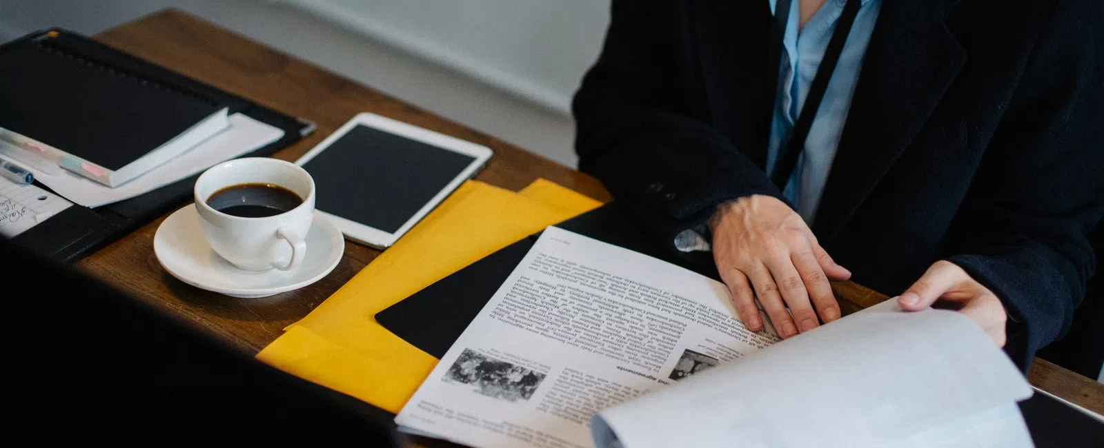 Person in business attire reading documents at desk with coffee, tablet, and folders.