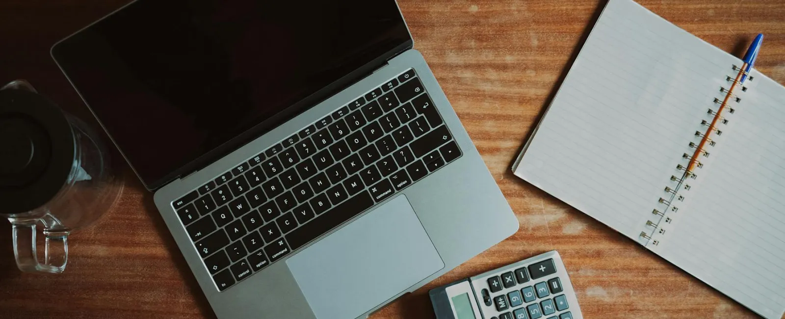 Overhead view of a laptop, calculator, open notebook with pen, and glass cup on wooden desk.
