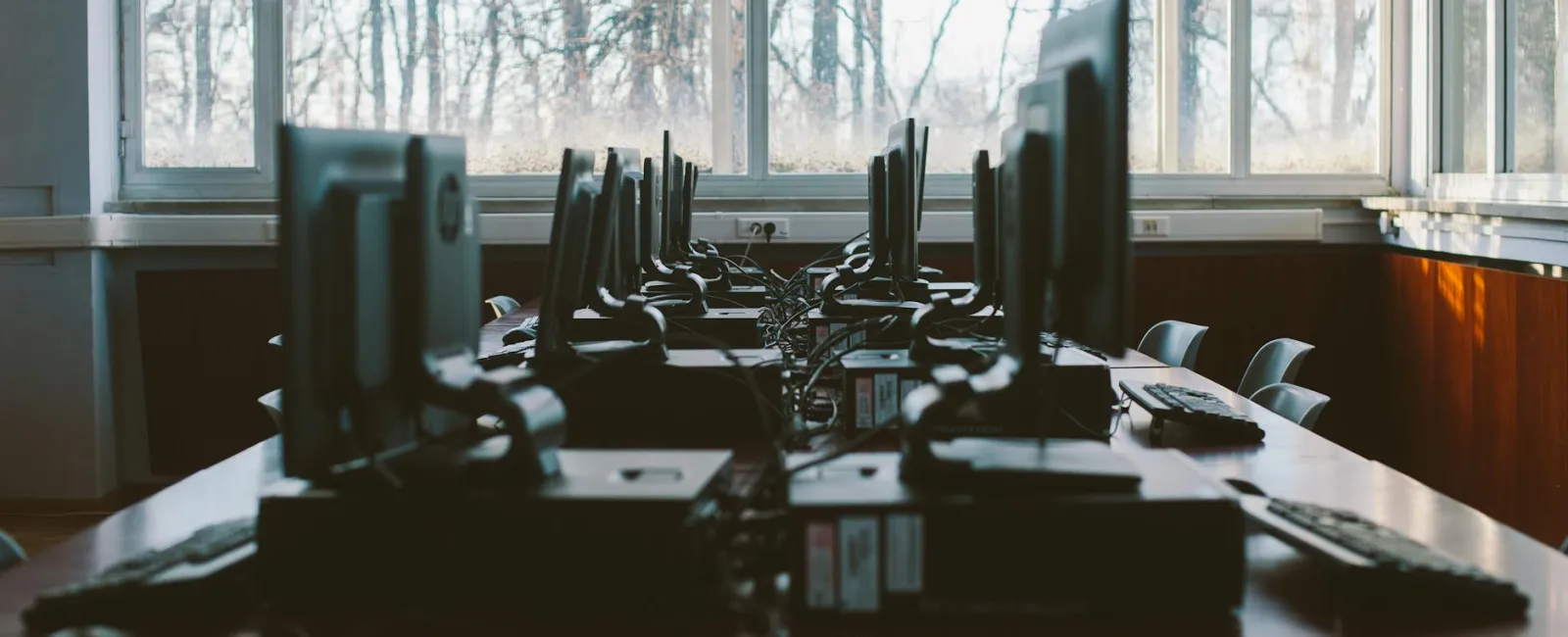 Empty computer lab with rows of desktop monitors and keyboards by large windows showing winter trees.