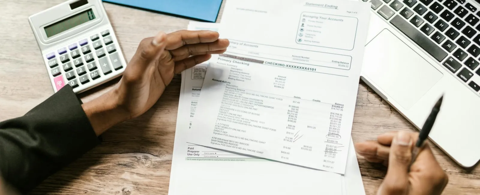 Person reviewing financial documents with calculator and laptop on wooden desk workspace