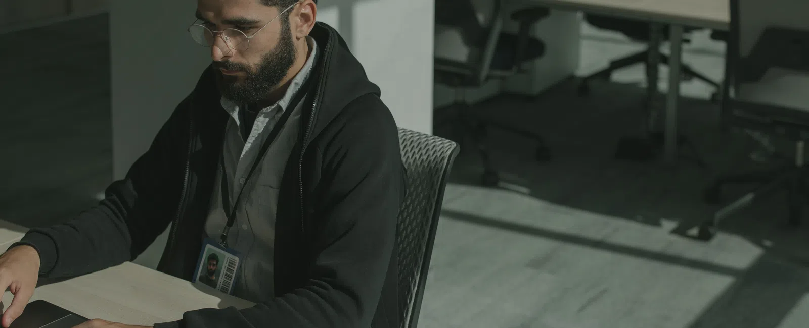 Man with curly hair and glasses working on a laptop in a modern, sunlit office space.