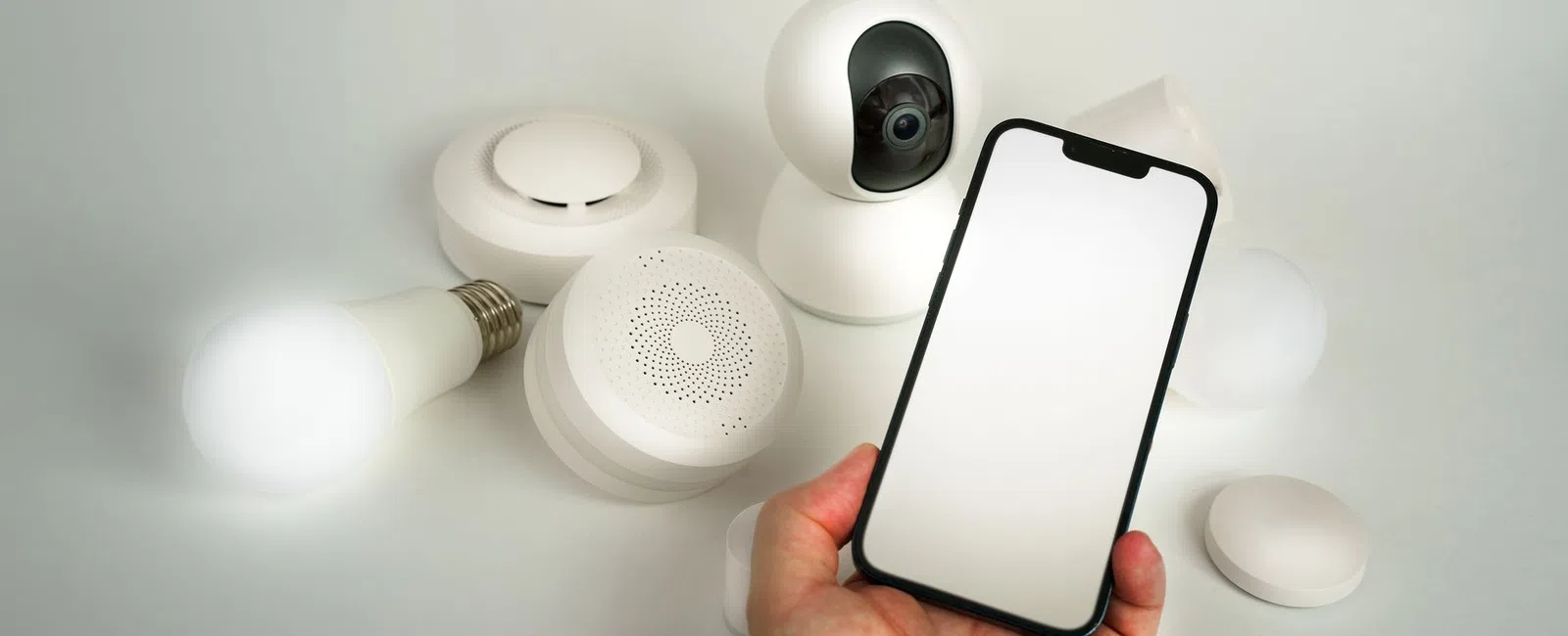 Hand holding smartphone with blank screen next to smart home devices including light bulb, camera, and sensors