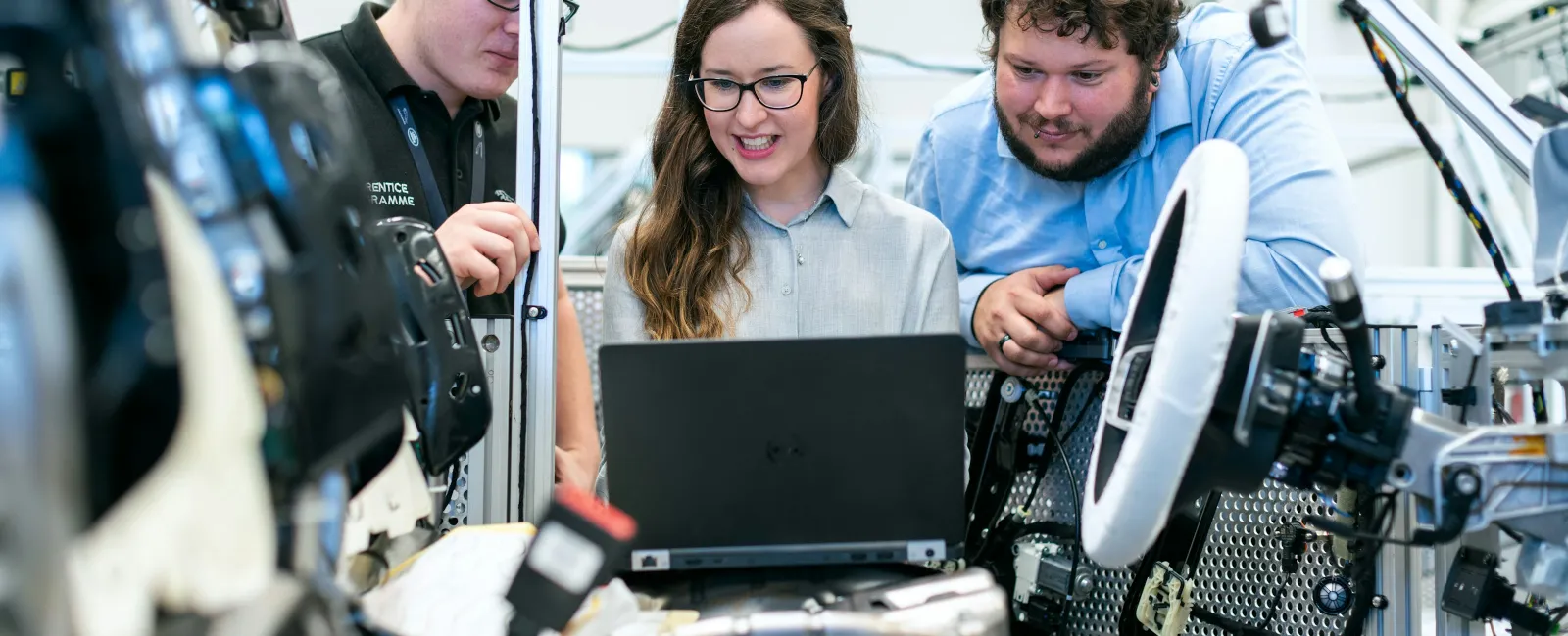 Three engineers collaborate on a laptop inside a vehicle assembly, focusing on automotive technology development.