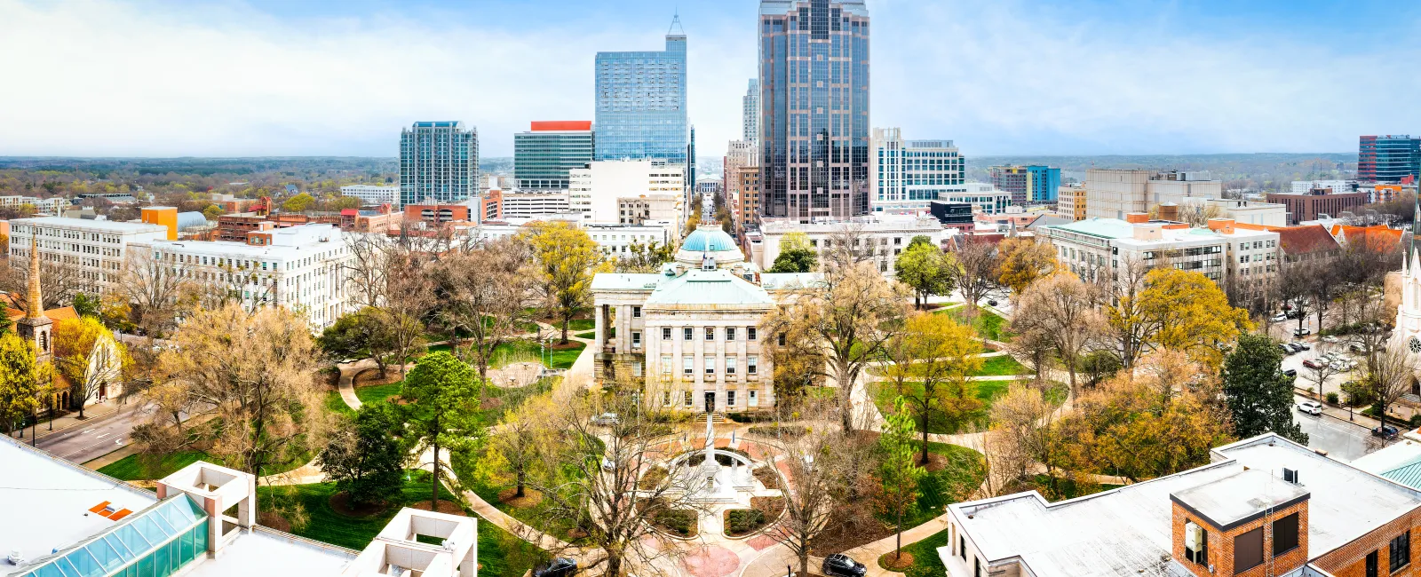 Aerial view of a city park with historic buildings, skyscrapers, and autumn trees under a blue sky.