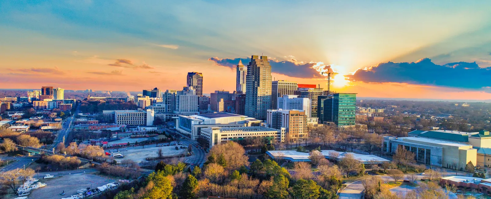 Aerial view of a city skyline at sunset with colorful sky and urban greenery in foreground.