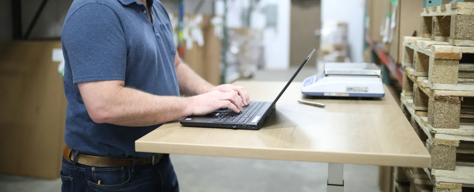 Man working on a laptop in a warehouse with shelves of pallets and packages around him.