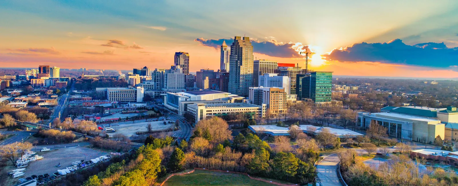 Sunset over a city skyline with tall buildings, green park in foreground, and a vibrant sky with clouds.