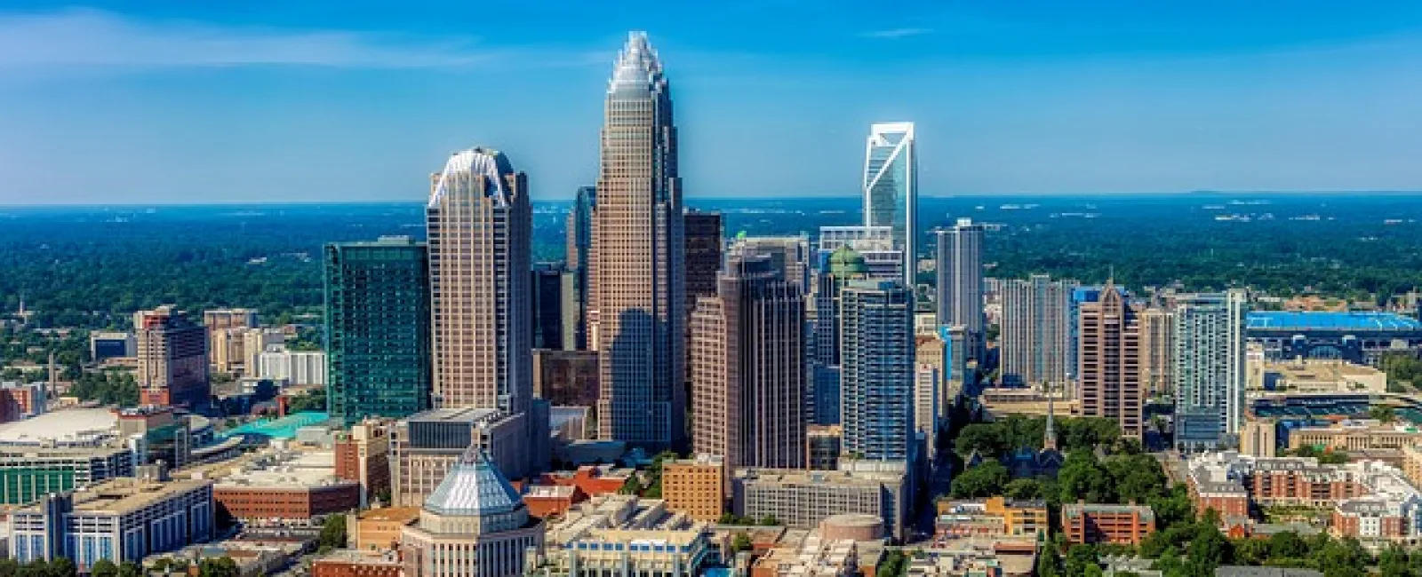 Aerial view of Charlotte city skyline with tall skyscrapers under a clear blue sky and surrounding greenery.