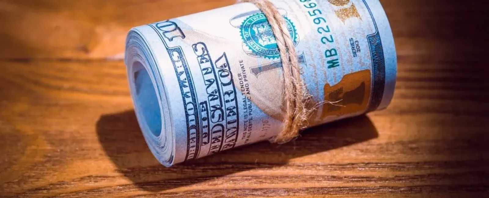 Rolled bundle of US 100 dollar bills tied with a string on a wooden surface.