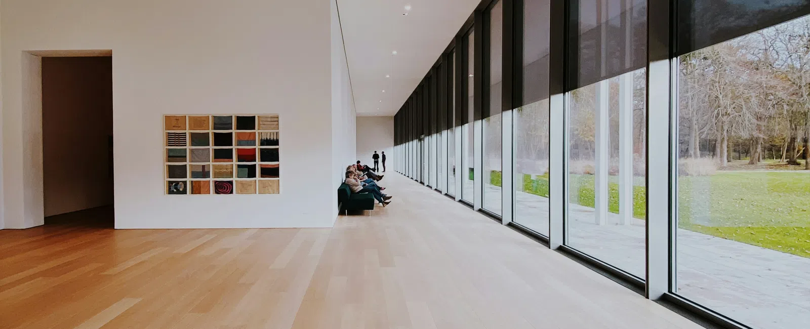 Modern art gallery with large windows, light wood floors, and visitors seated along white walls.