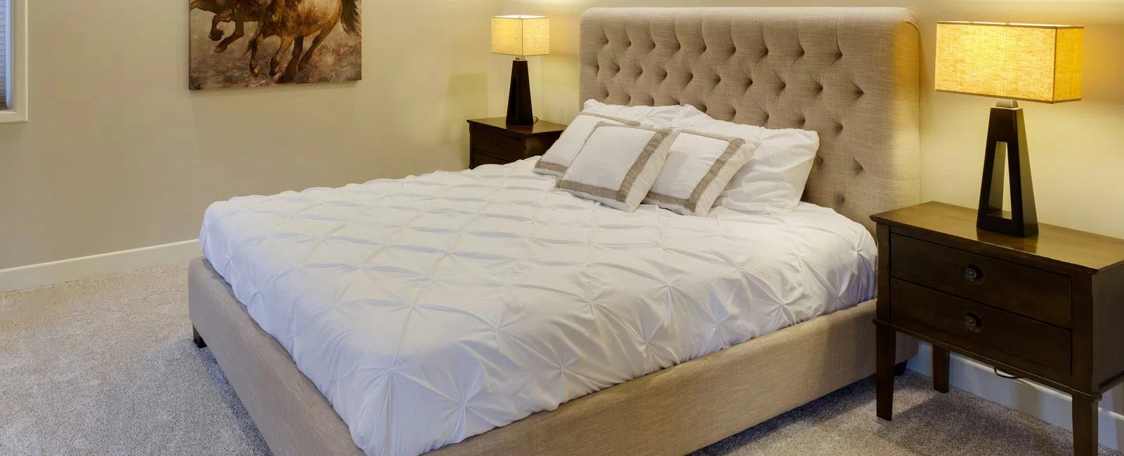 Cozy beige bedroom with tufted bed, white bedding, two nightstands, lamps, and horse painting on wall.