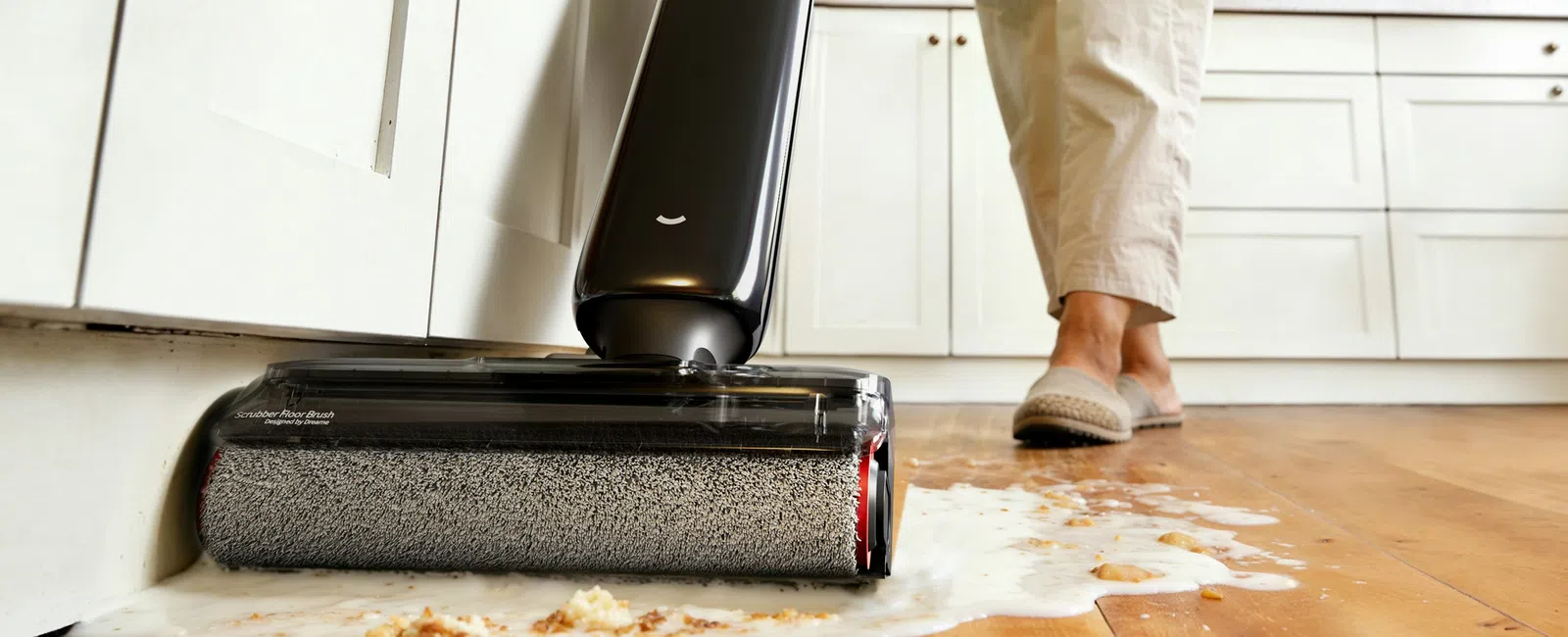 Person using an electric floor cleaner to mop spilled juice and crumbs off a hardwood kitchen floor.