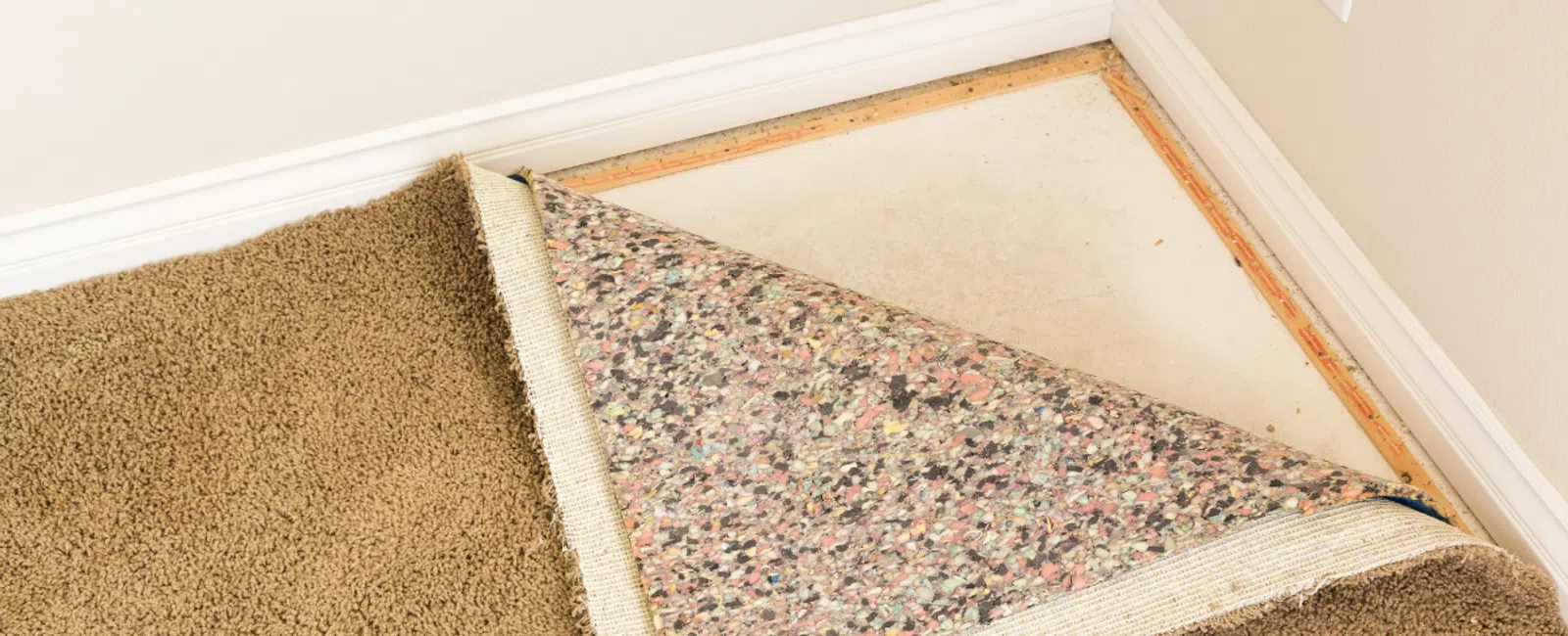 Beige carpet partially pulled back revealing carpet padding and bare floor in a corner room with white baseboards.