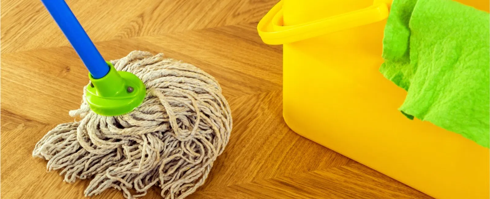 Wet mop head and yellow cleaning bucket with green cloth on wooden parquet floor.