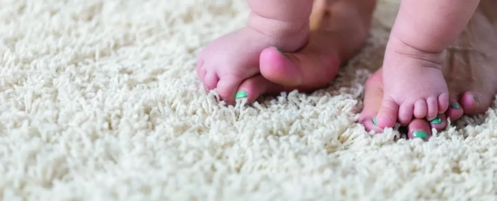 Close-up of baby feet standing on adult feet with green painted toenails on a soft cream rug.