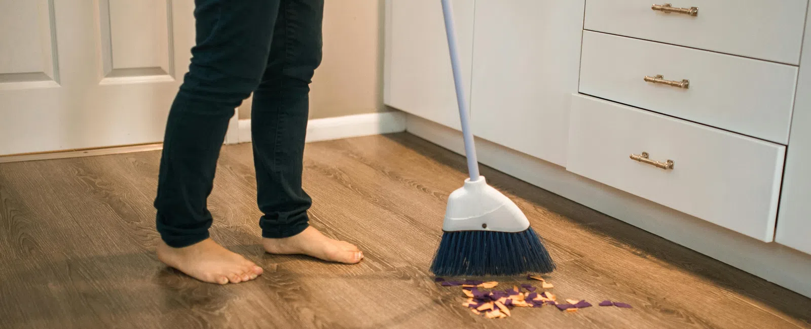 Person sweeping trash on wooden kitchen floor barefoot near white cabinets and drawers.
