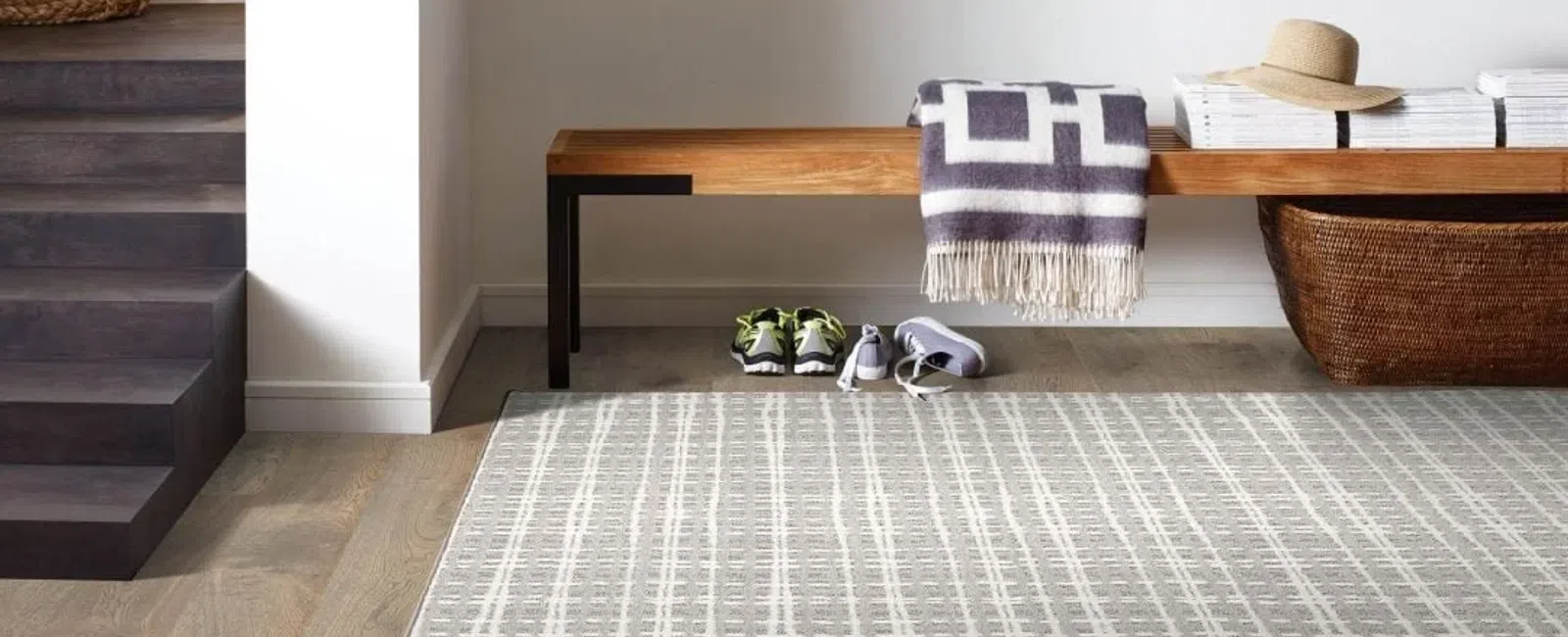 Modern entryway with wooden bench, patterned rug, woven basket, folded blanket, shoes, and hat under soft natural light
