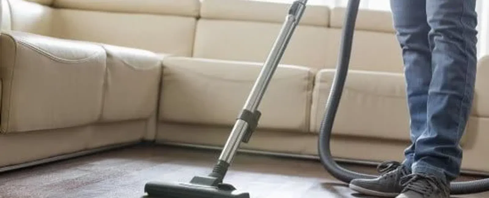 Person vacuuming a wooden floor next to a beige sectional sofa in a bright living room.