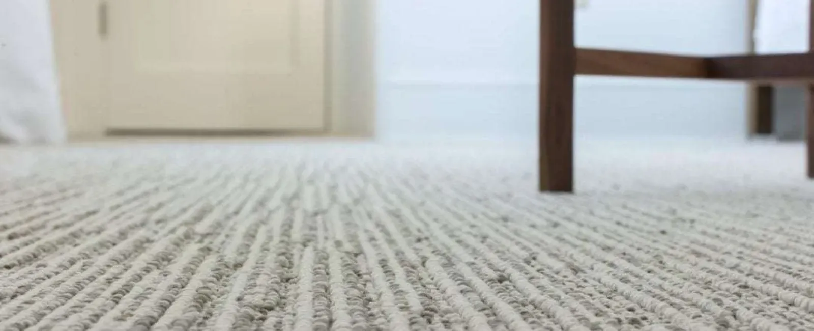 Close-up of textured beige carpet with wooden chair legs and white door in background inside bright room