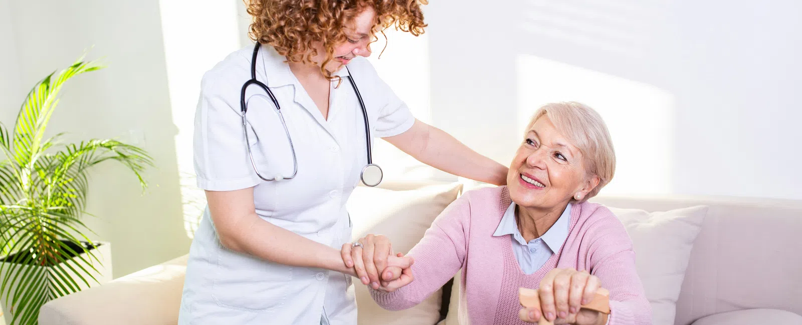 Nurse assisting smiling elderly woman with walking cane in a bright living room setting.