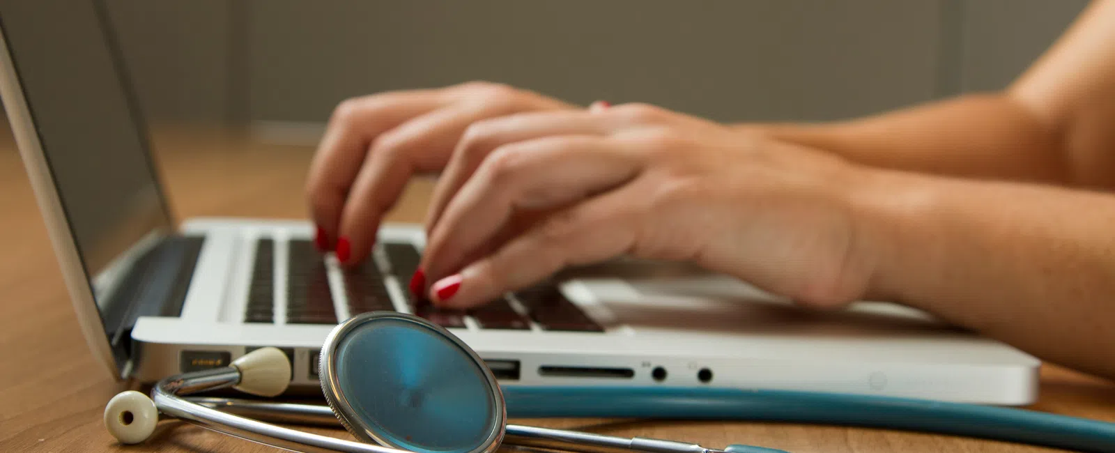 Hands typing on a laptop keyboard with a stethoscope resting on the wooden table beside it showcasing healthcare IT at work.