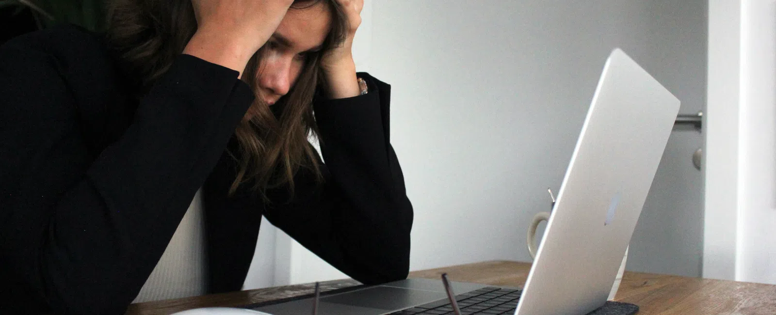 Stressed woman holding head sitting at wooden desk with laptop, phone, glasses, and mouse, working indoors.