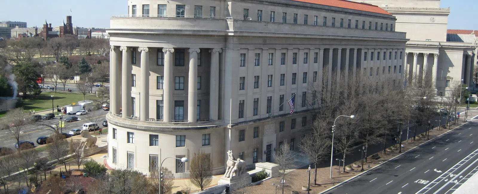 Historic government building with columns and red roof alongside a widHome of the feHome of the federal trade commission (FTC), the historic Apex building has columns and red roof beside a wide street. deral trade commission (FTC), Apex building with columns and red roof beside a wide street. e street with sparse traffic under a blue sky.