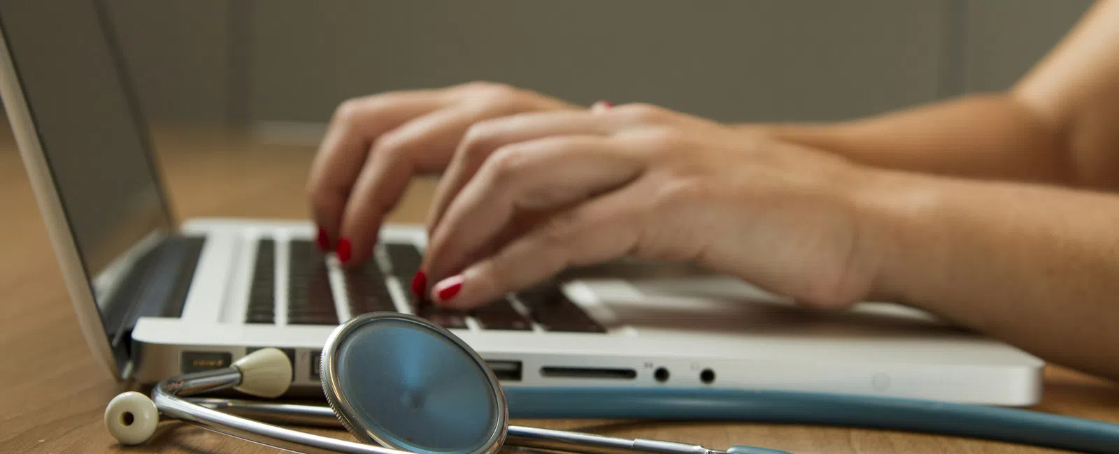 Hands typing on a laptop keyboard with a stethoscope placed on the wooden desk nearby, indicating medical work.