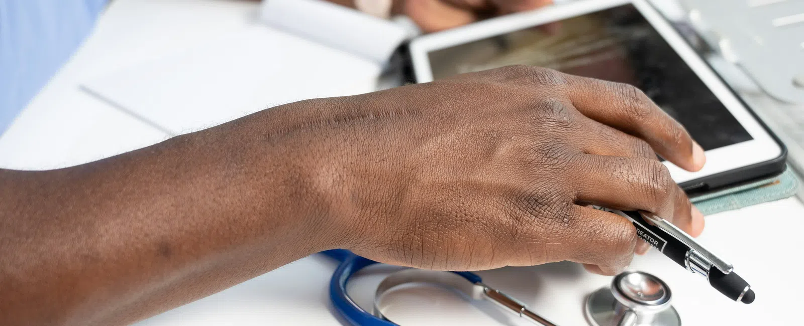 Close-up of a doctor's hand using a tablet with a stethoscope on the desk, representing medical care technology.
