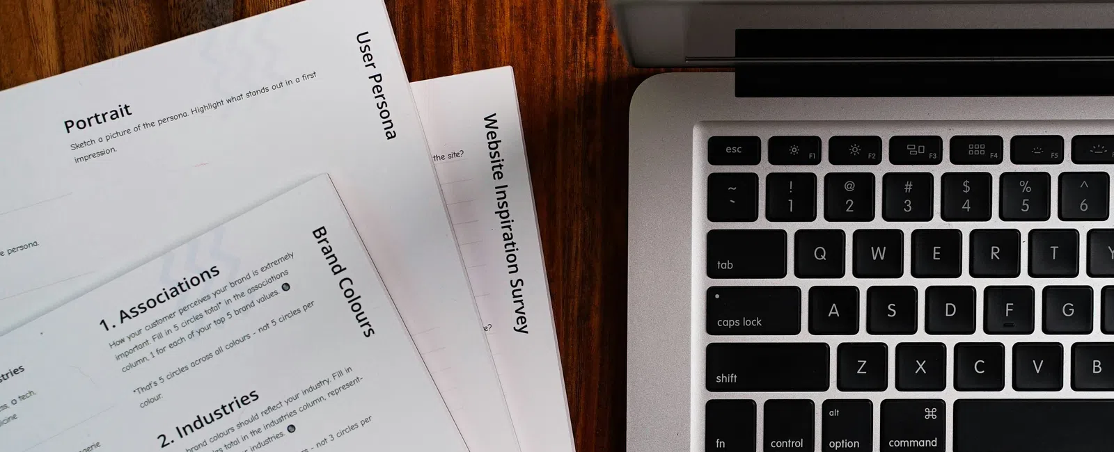 Close-up of a laptop keyboard beside printed user persona and brand color survey documents on a wooden desk
