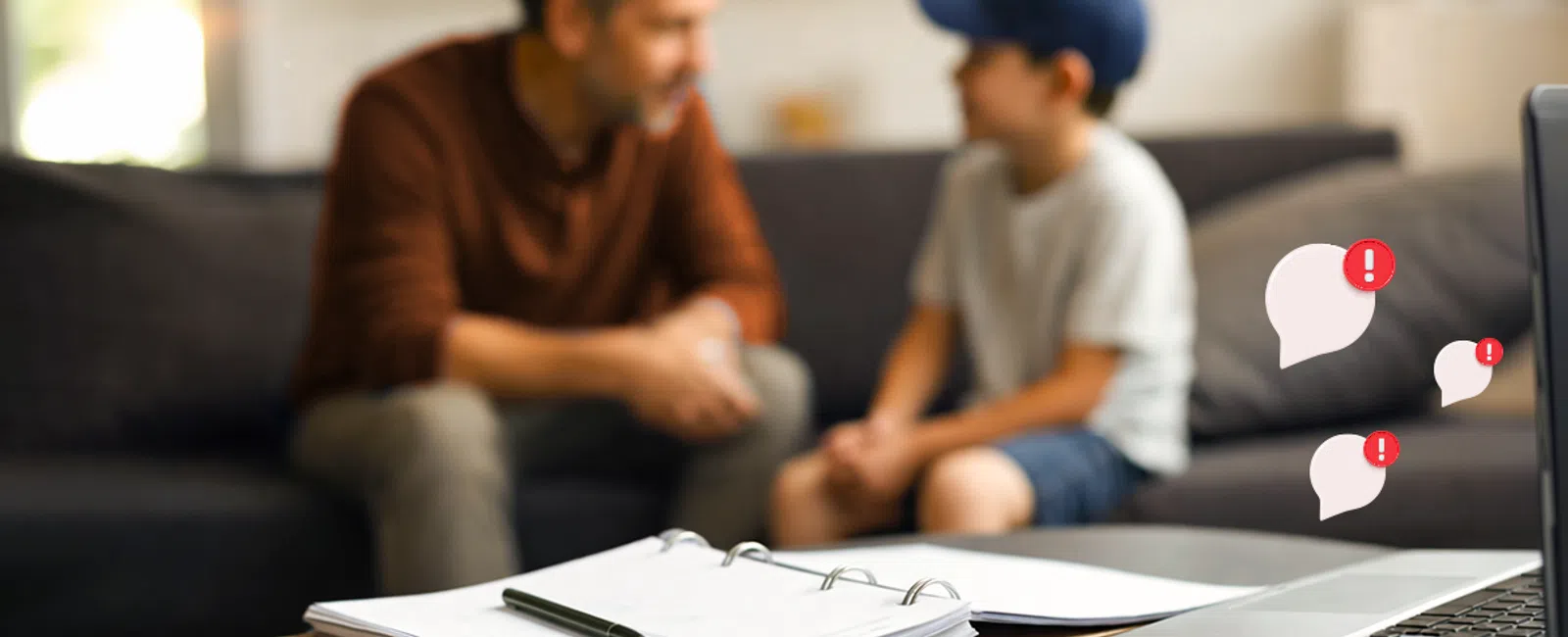 Father and son talking on couch with laptop, notebook, and smartphone on table in foreground