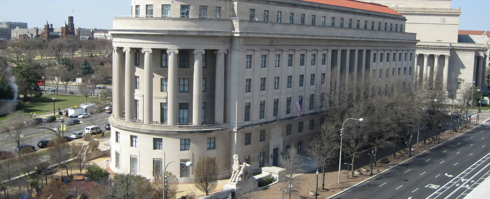Historic government building with columns and red roof alongside a widHome of the feHome of the federal trade commission (FTC), the historic Apex building has columns and red roof beside a wide street. deral trade commission (FTC), Apex building with columns and red roof beside a wide street. e street with sparse traffic under a blue sky.