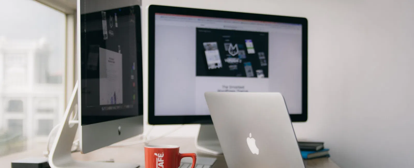 Workstation setup with Apple laptop, iMac, Nescafé red mug, and dual monitors on a wooden desk near window.